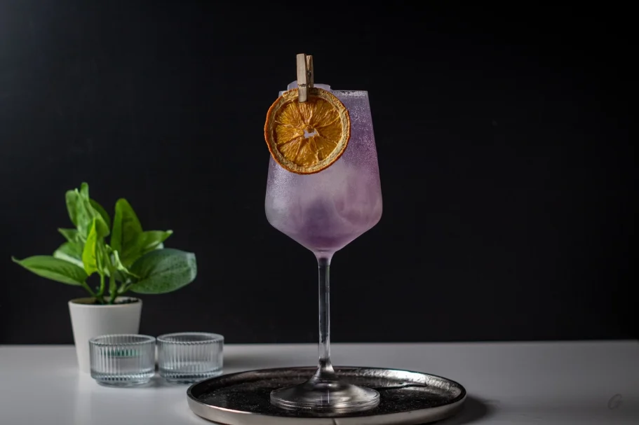 Purple Kiss Cocktail violet avec une garniture de patate douce sur un plateau, accompagné d'une plante.