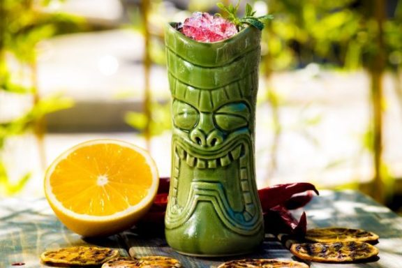 Zombie Tasse tiki verte avec un cocktail décoré, tranches d'orange et biscuits en arrière-plan.