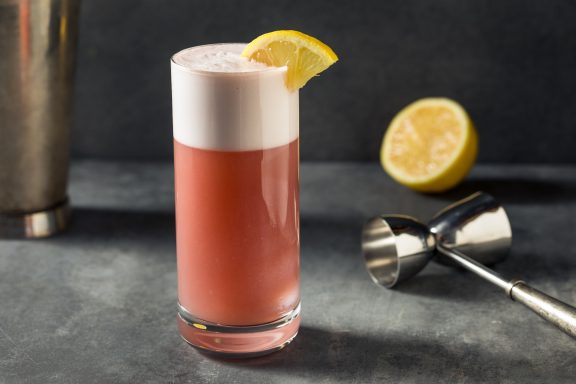 Chicago Fizz Cocktail rose avec mousse, garnie d'une tranche de citron, sur une surface sombre.
